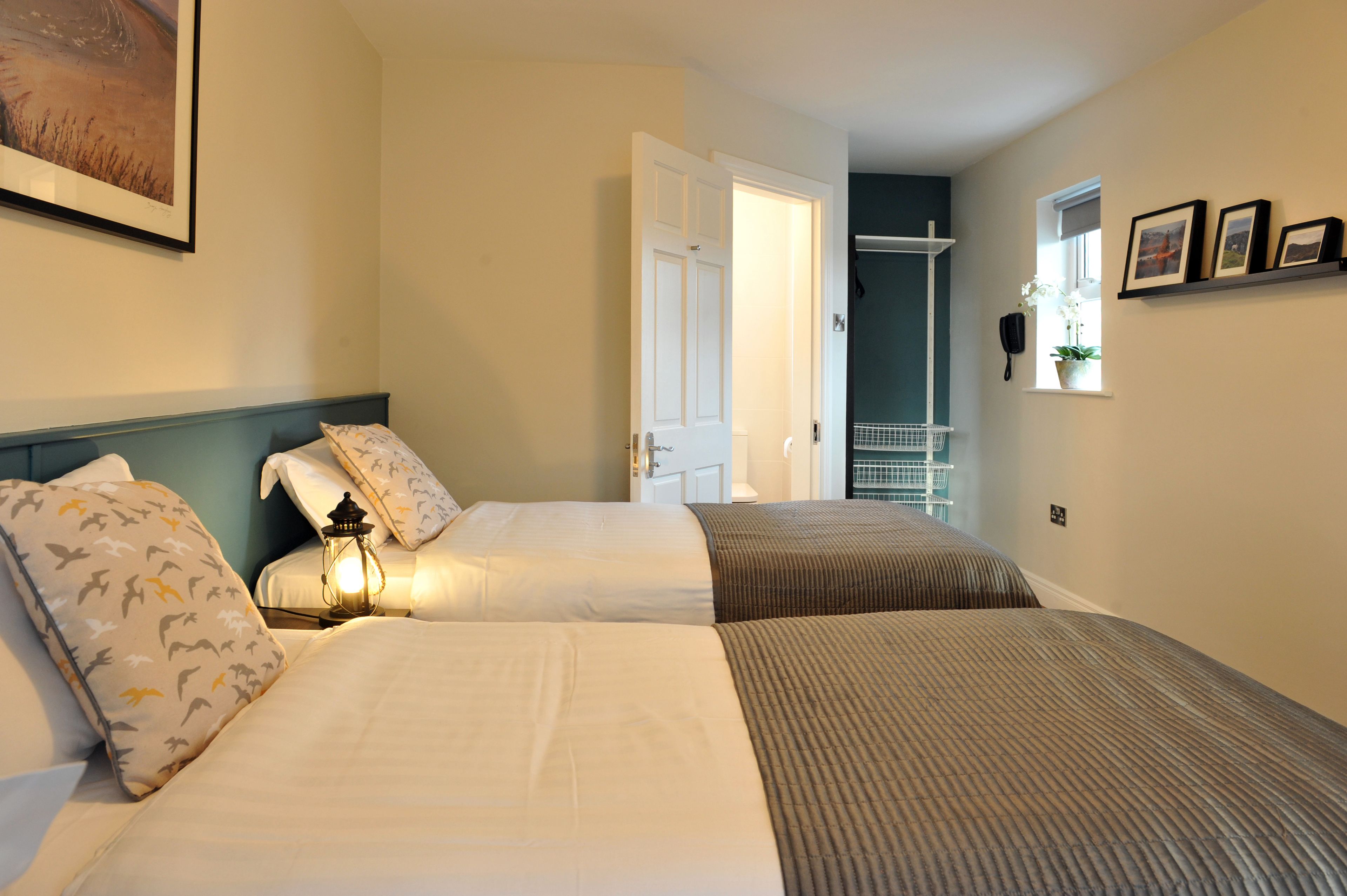 A hotel room with two beds, a nightstand with a lamp, a door leading to a balcony, and a window with a plant.