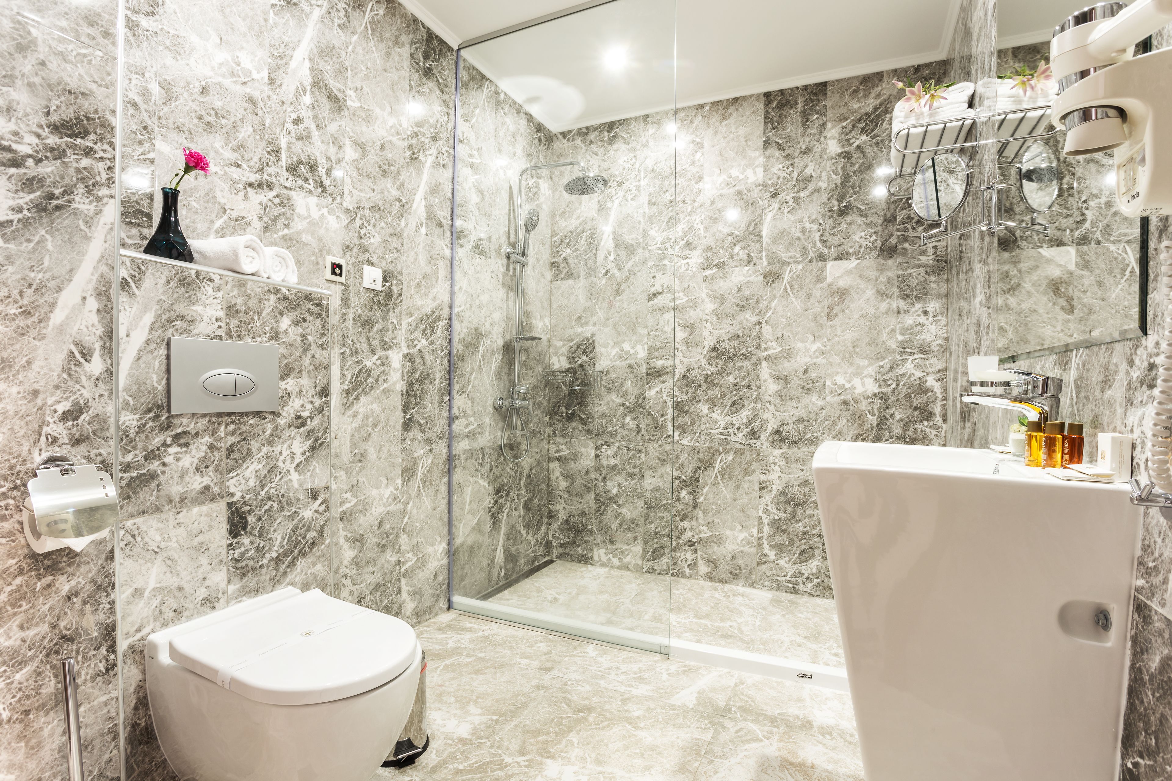 A modern bathroom with a glass-enclosed shower, marble walls, a white toilet, and a sink with toiletries.
