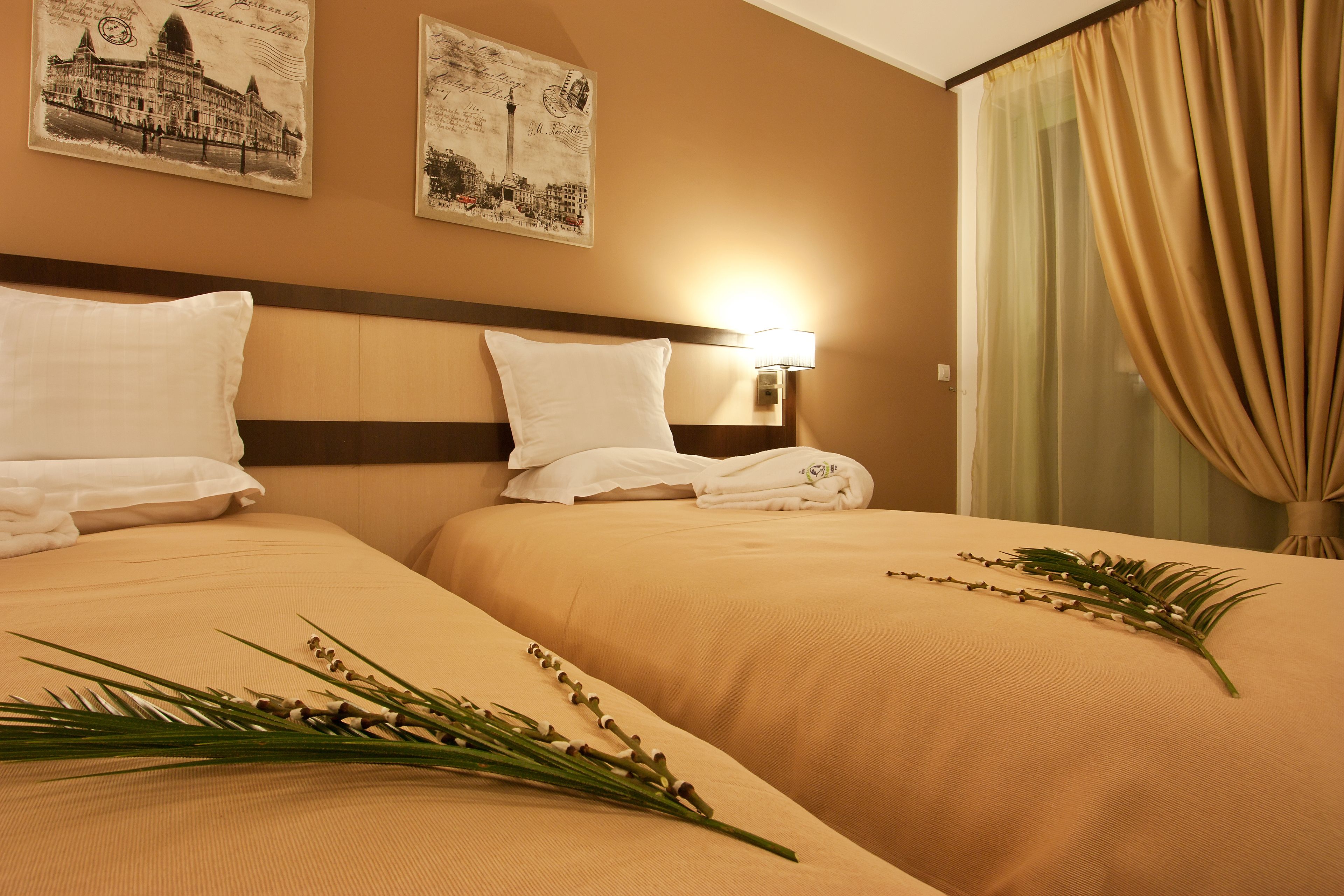 Habitación de hotel con dos camas, ropa de cama color crema, una mesita de noche con lámpara y dos cuadros en la pared.