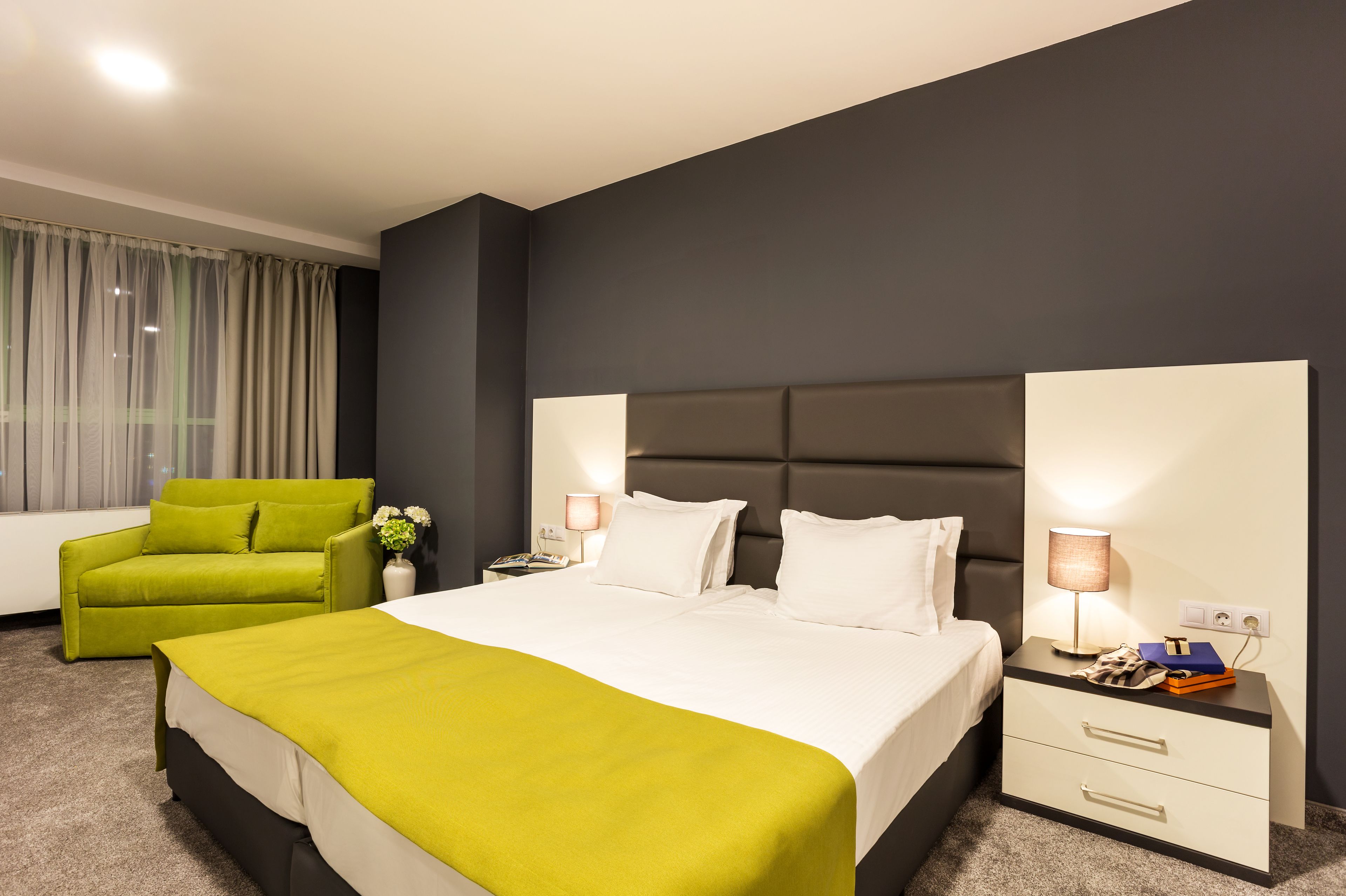 Una habitación de hotel con una cama, un sillón verde, una mesita de noche con lámpara y un pequeño mueble auxiliar.