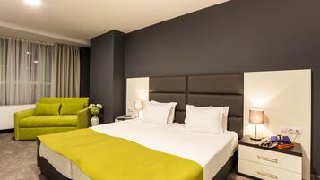 Una habitación de hotel con una cama, un sillón verde, una mesita de noche con lámpara y un pequeño mueble auxiliar.