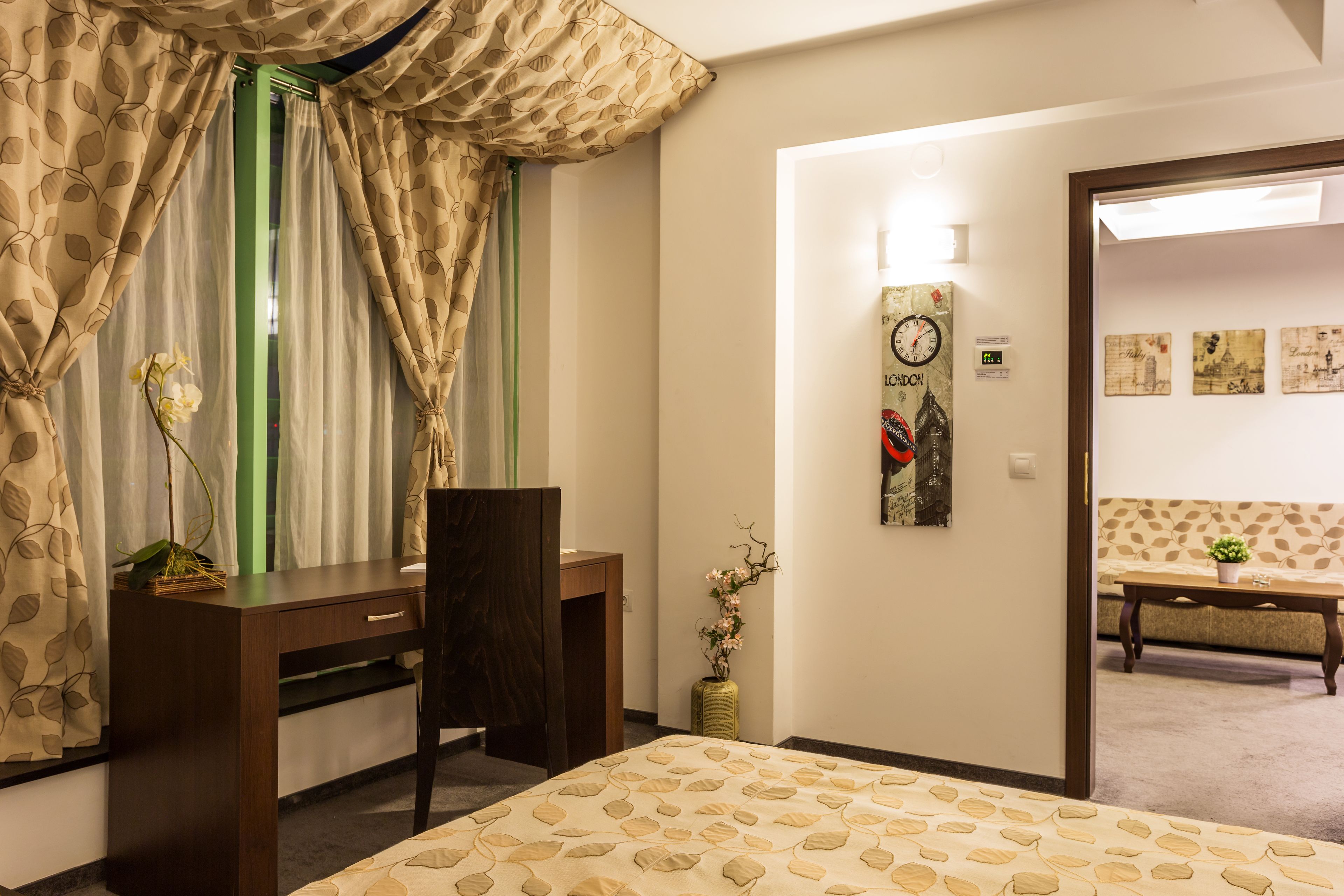 Quarto de hotel com cama, escrivaninha, cadeira, vaso de flores e relógio na parede.