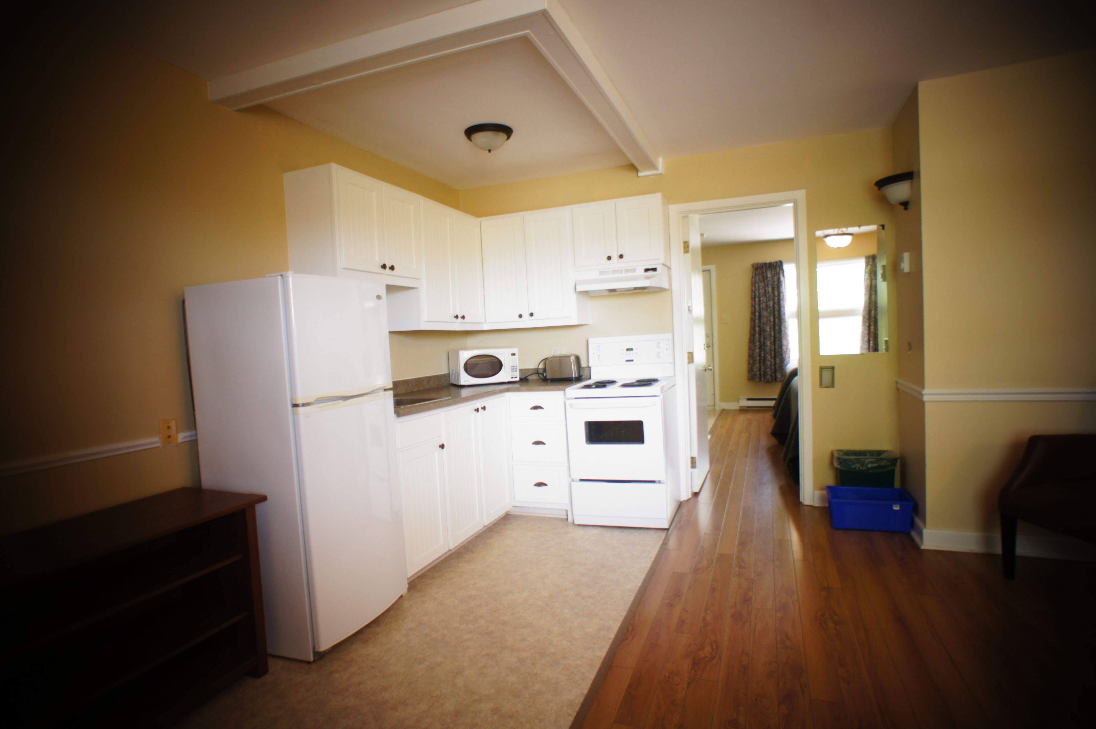 A compact kitchen with white cabinets, a refrigerator, microwave, and stove. A wooden bench is against the wall, and a blue storage box is on the floor.