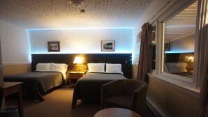 A hotel room with two beds, a nightstand, a lamp, a chair, and a small table. - Silverwood Inn & Suites (Fredericton)