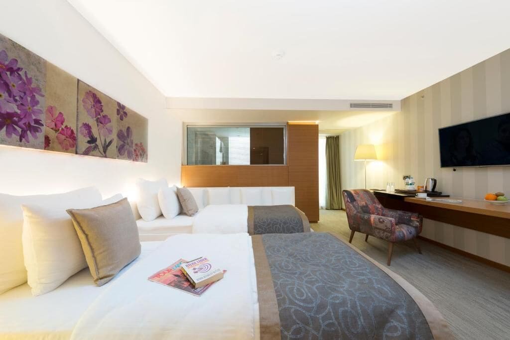 A hotel room with two beds, a desk, a chair, a TV, and a large floral artwork on the wall.