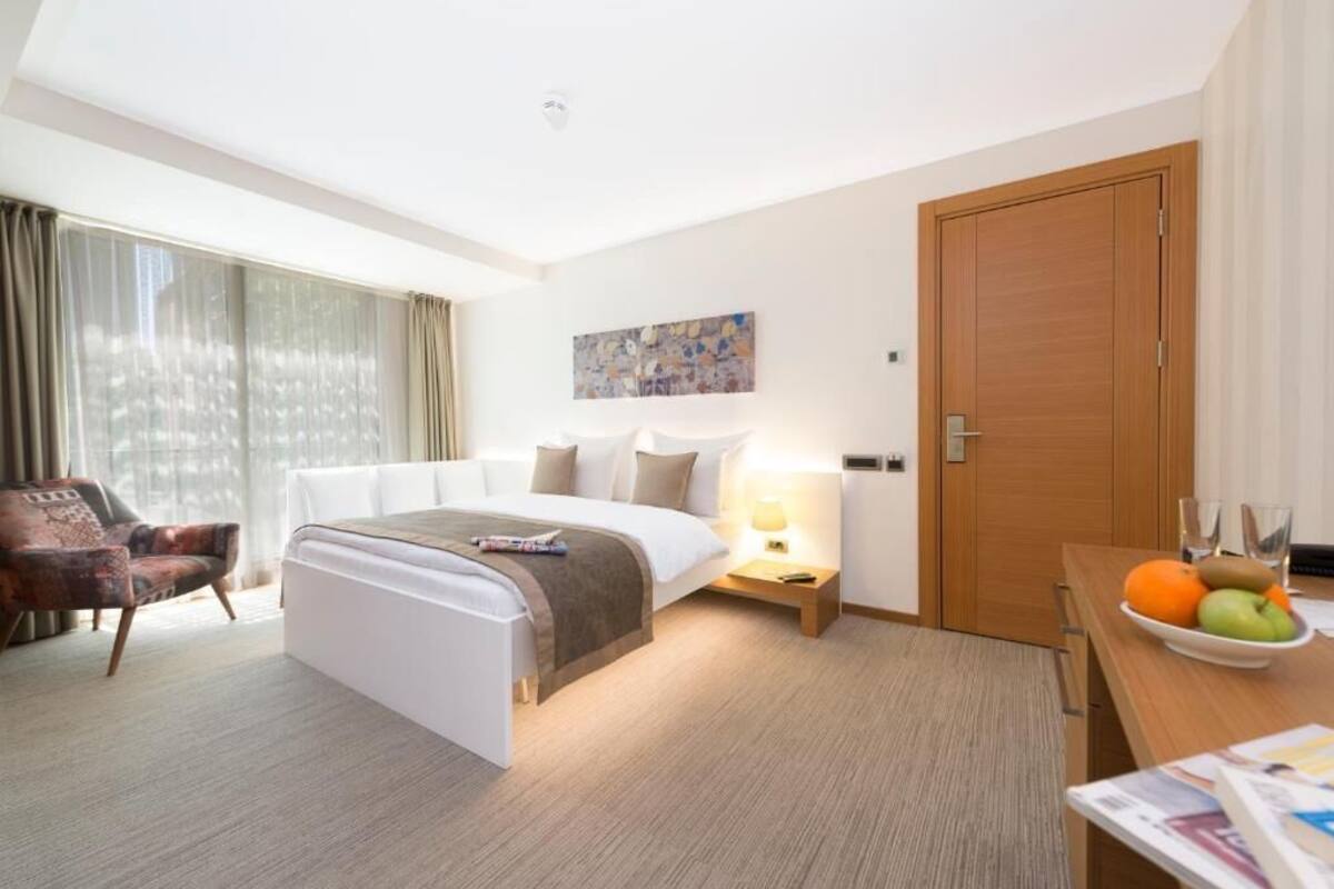 A hotel room with a bed, bedside table, lamp, chair, and a view of the outdoors through the window.