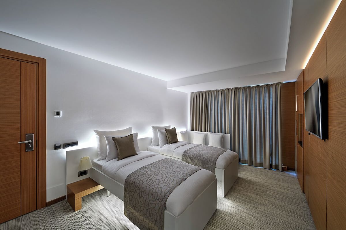 A modern hotel room with two single beds, a wooden wardrobe, a flat-screen TV, and a large window with curtains.