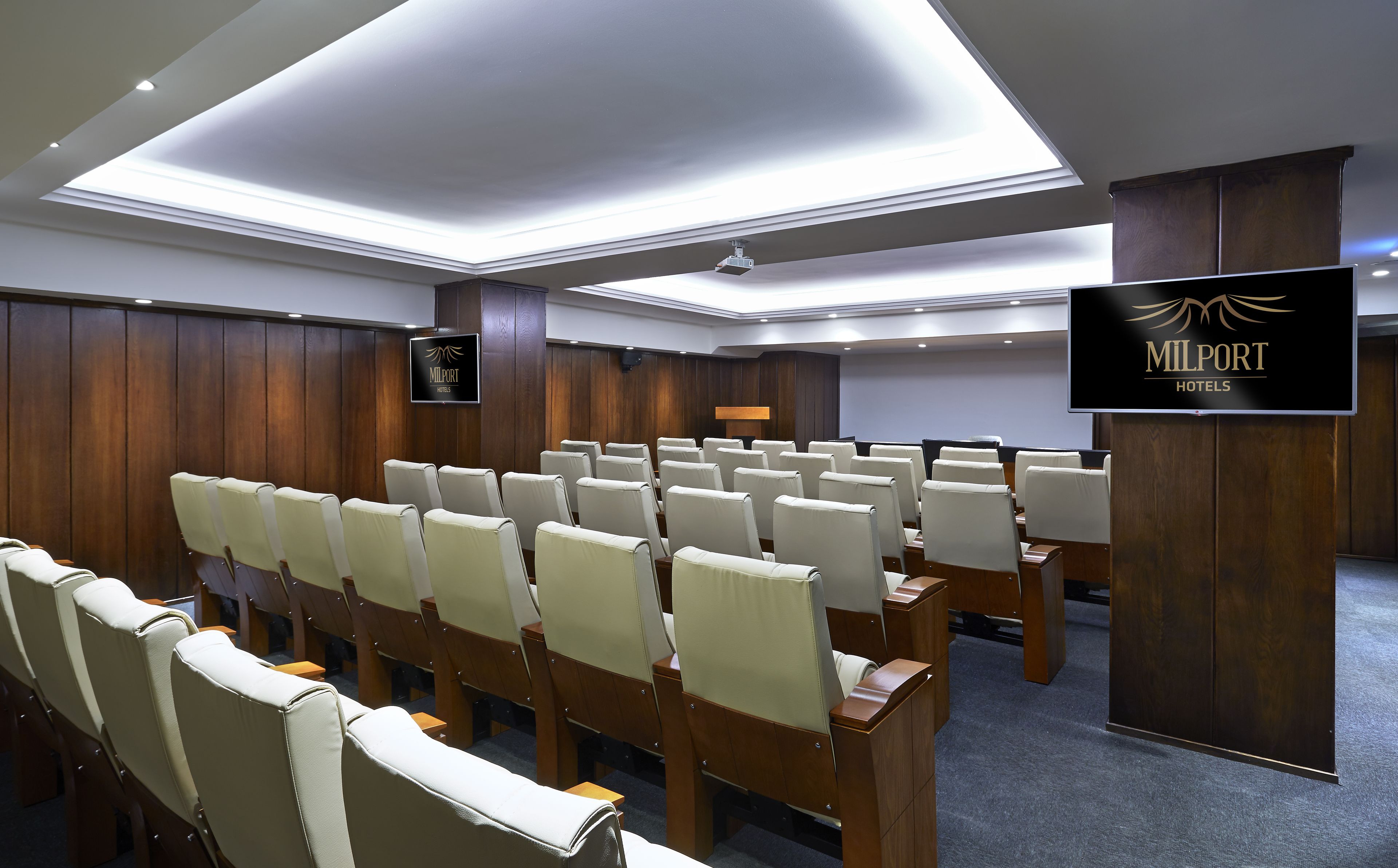 Een vergaderzaal met rijen witte stoelen, een spreekgestalte en een bord met Milport Hotels.