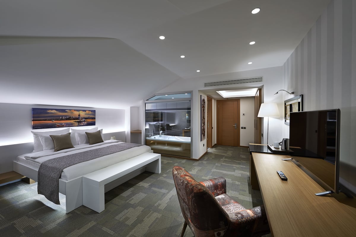 A modern hotel room with a large bed, a desk, a chair, a television, and a bathroom with a glass shower.