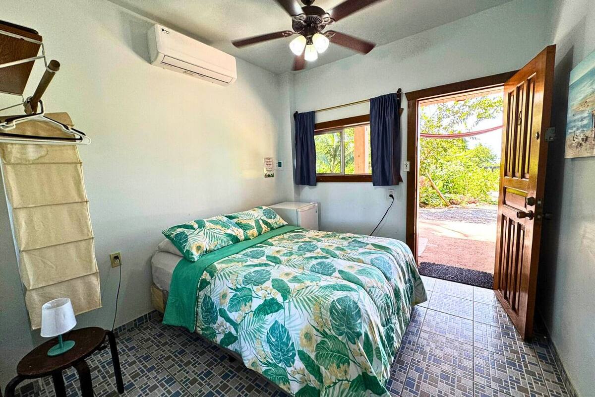 A bedroom with a bed, a ceiling fan, an air conditioner, a window with curtains, a door, and a small table with a lamp.