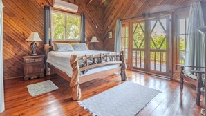 Une chambre dans une cabane en bois, avec un lit, deux tables de chevet, un ventilateur de plafond et de grandes fenêtres.