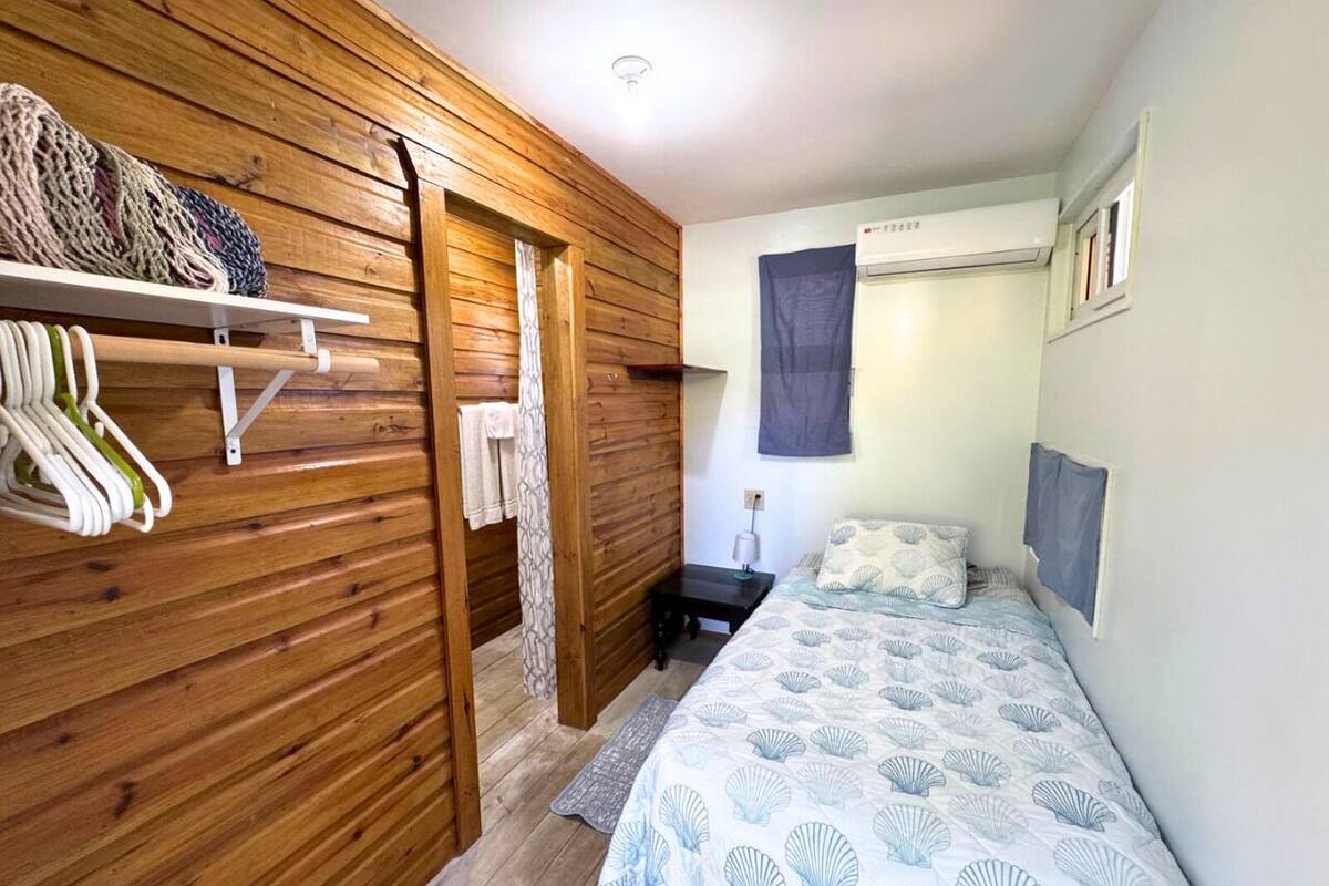 A bedroom with a wooden wardrobe, a bed with a seashell pattern bedspread, a nightstand, and a wall-mounted air conditioner.