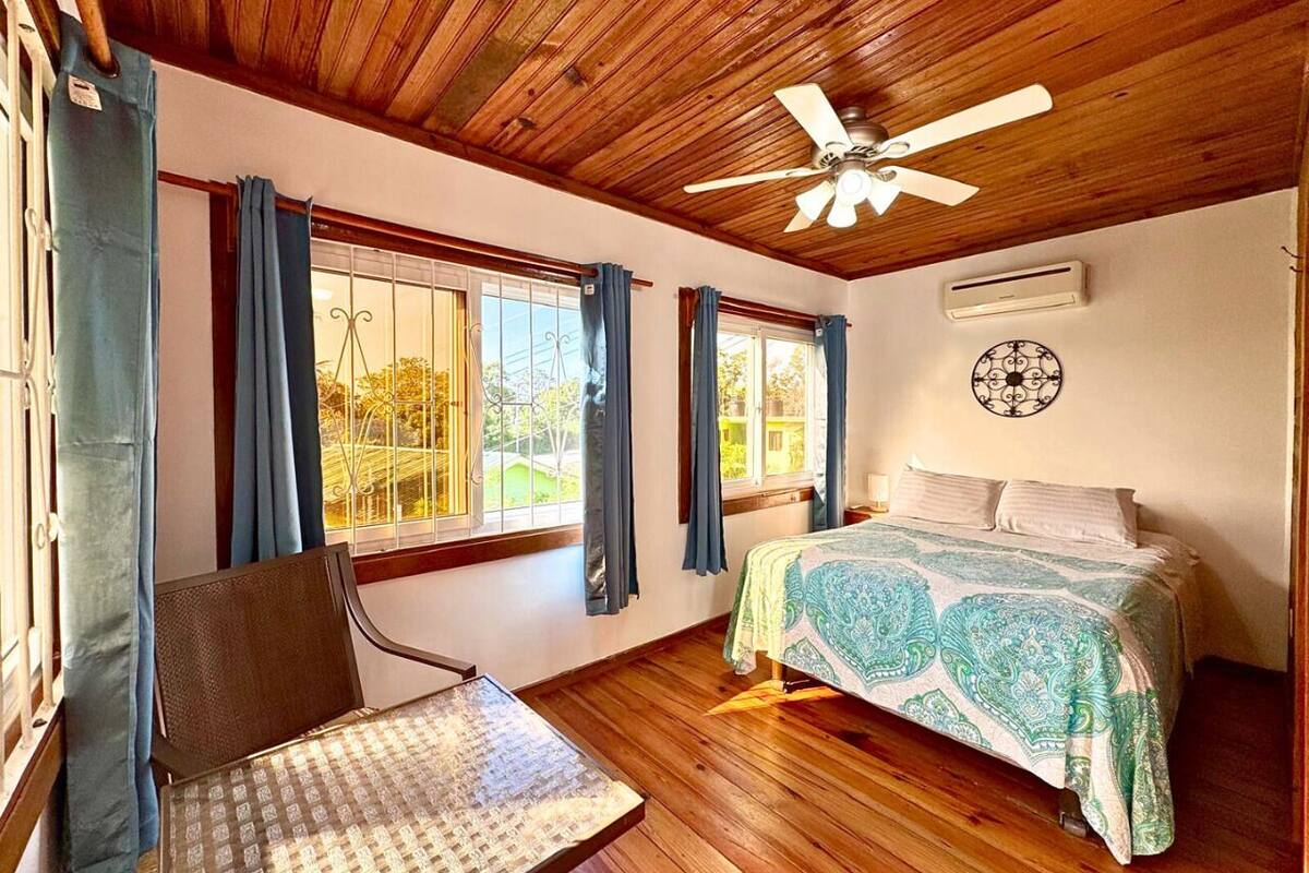 A bedroom with a wooden ceiling, a bed with patterned bedding, a chair, and a window with curtains.