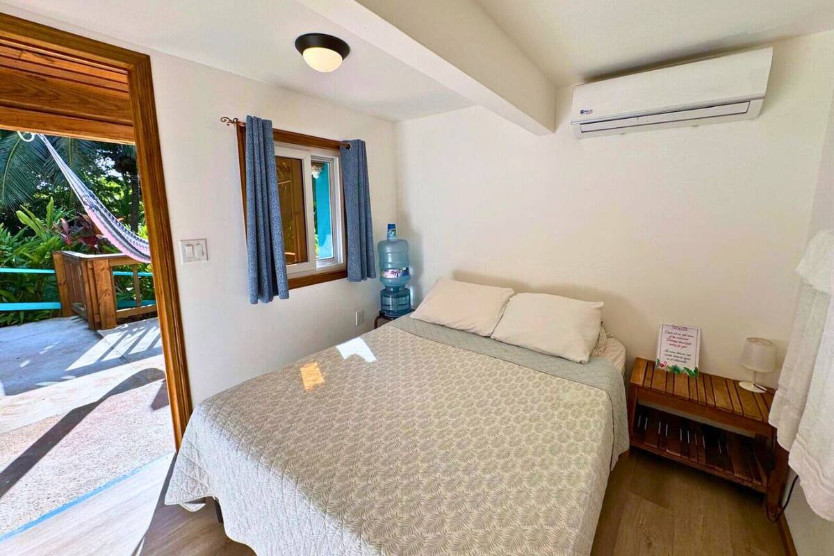 A bedroom with a bed, a water dispenser, a small table with a lamp, and a view of the outdoors.