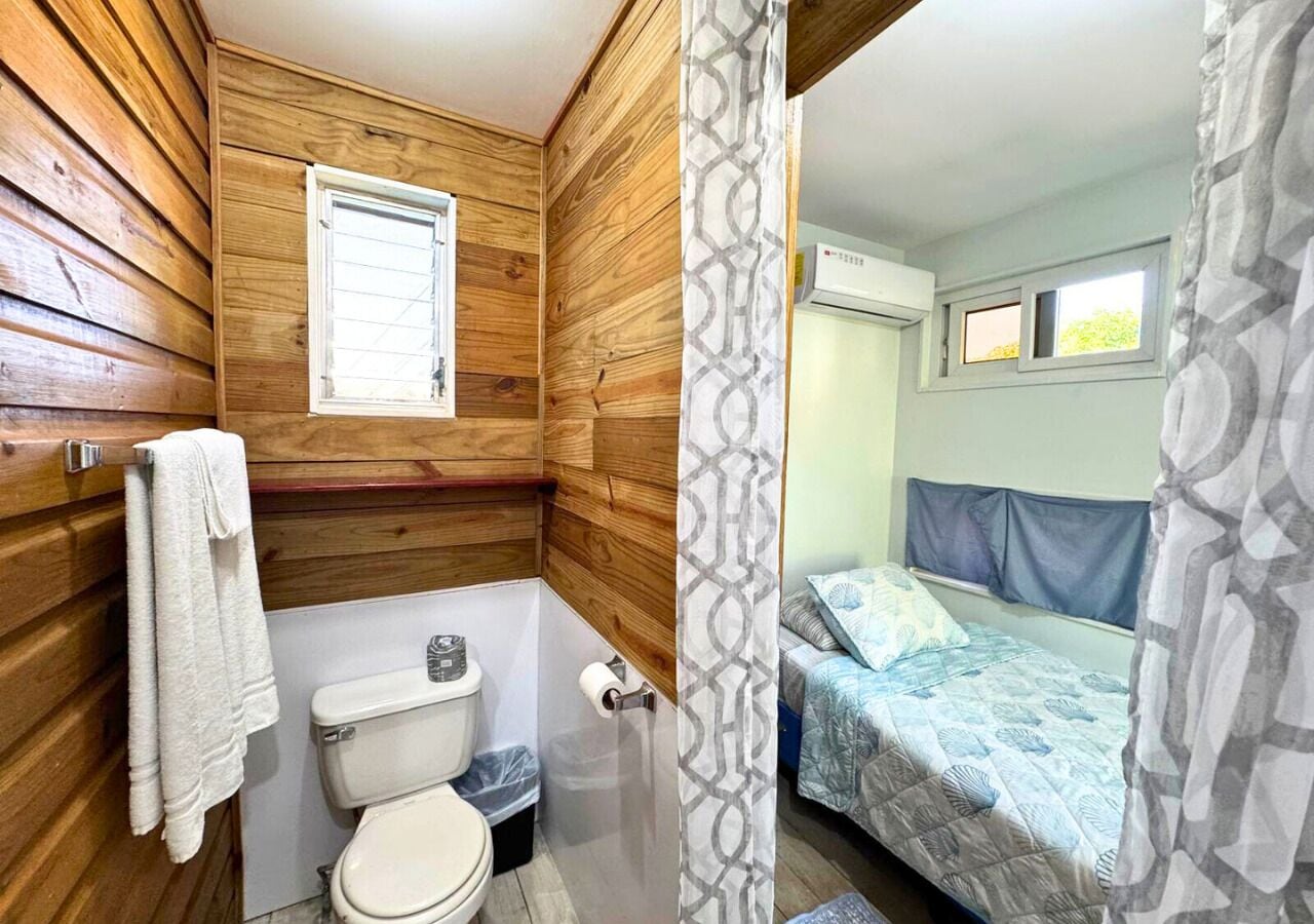 A small, compact room with wooden walls, a toilet, a shower, and a bed with a patterned bedspread.