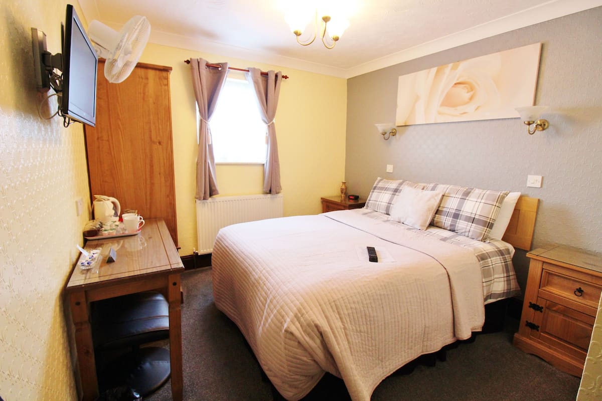 A bedroom with a bed, a desk, a chair, a nightstand, a lamp, and a window with curtains.