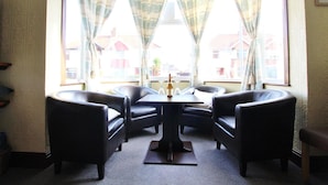 A room with a round table and four black leather chairs, large windows with curtains, and a view of houses outside. - Maluth Lodge (Great Yarmouth)