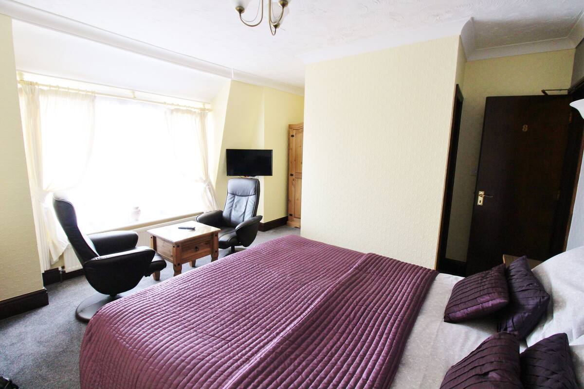 A hotel room with a large bed, a desk, a chair, a television, and a wardrobe.