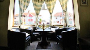 A dining area with a table and four chairs, large windows with curtains, and a view of buildings outside. - Maluth Lodge (Great Yarmouth)