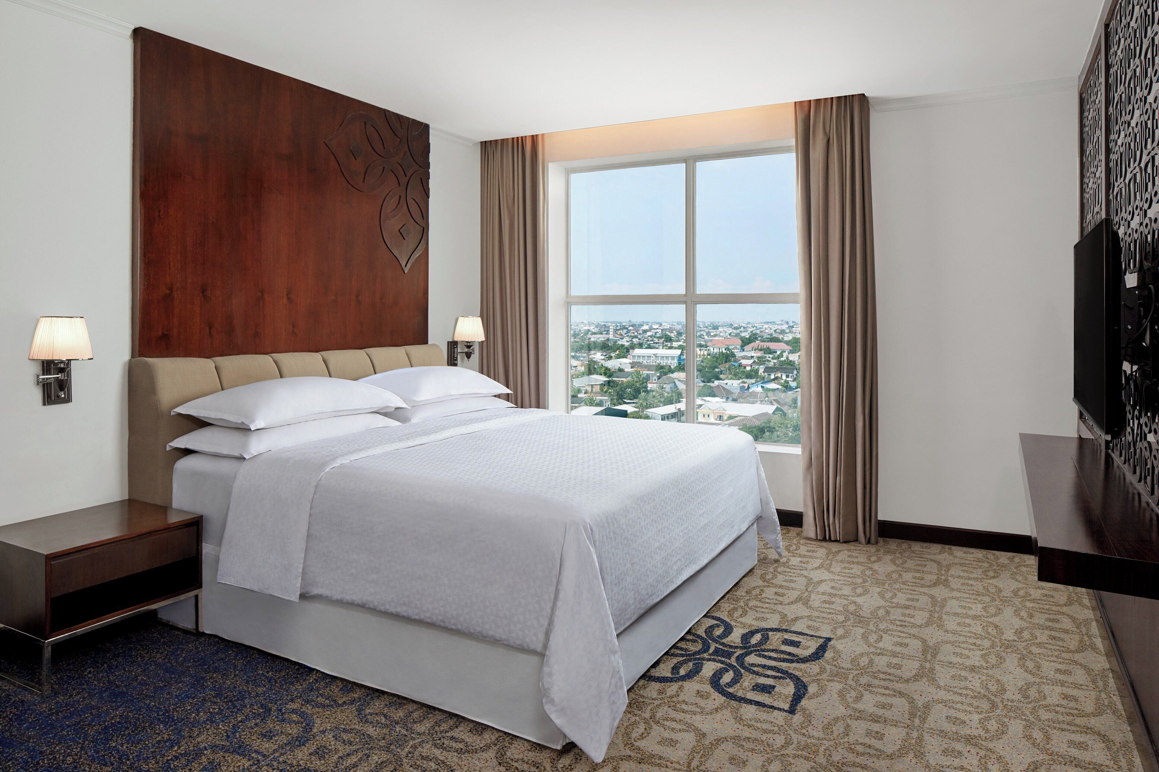 Habitación de hotel con una cama grande, dos mesitas de noche, un televisor y una ventana con vistas a la ciudad.