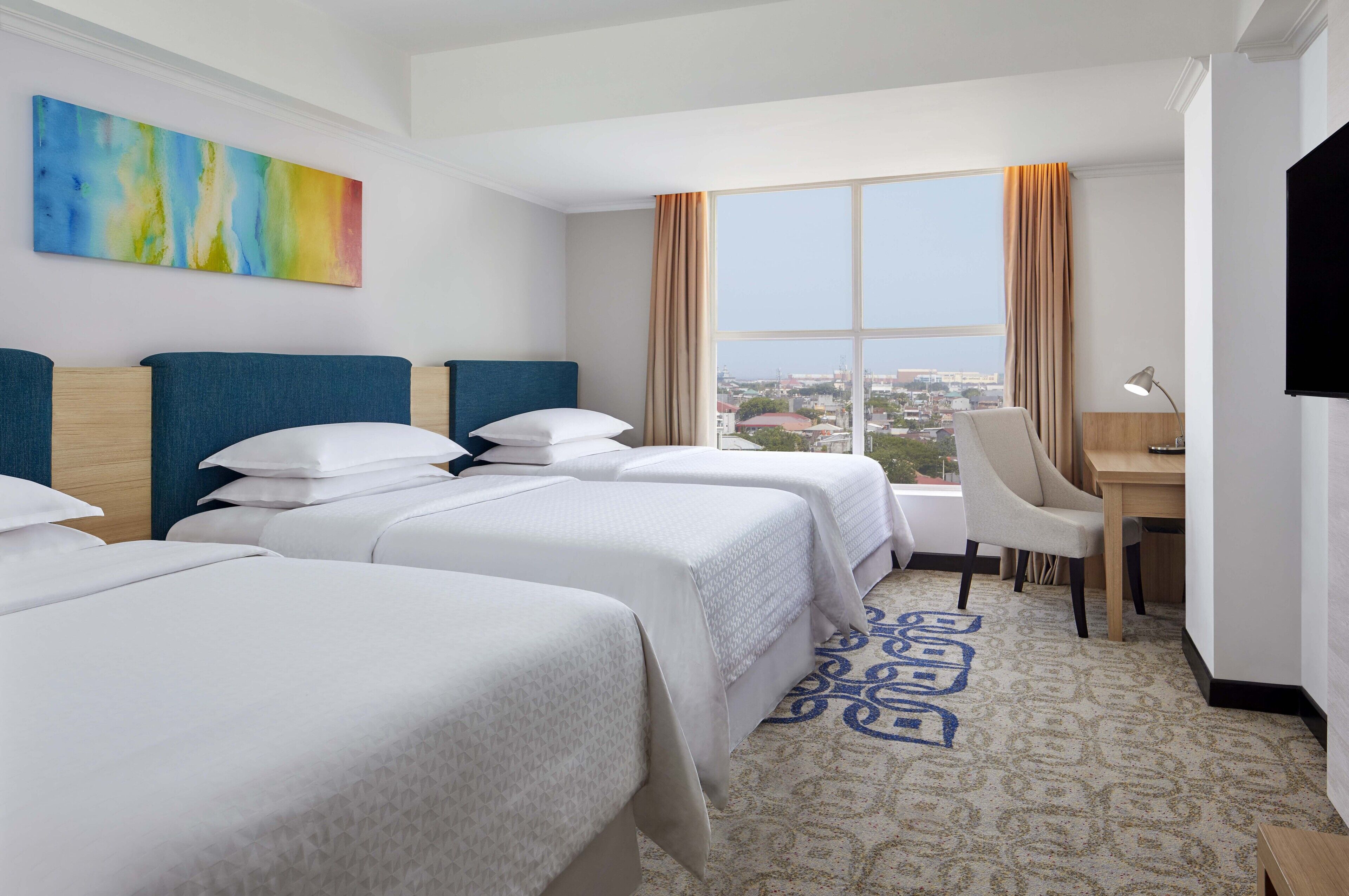 A hotel room with three beds, a desk, a chair, a TV, and a large window with a city view.
