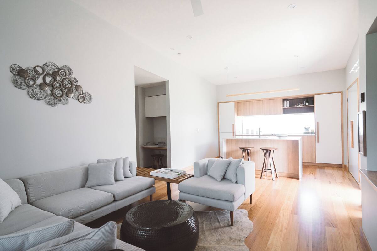 A modern living room with a grey sofa, a smaller armchair, and a coffee table. A kitchen is visible in the background with wooden cabinets and a bar stool.