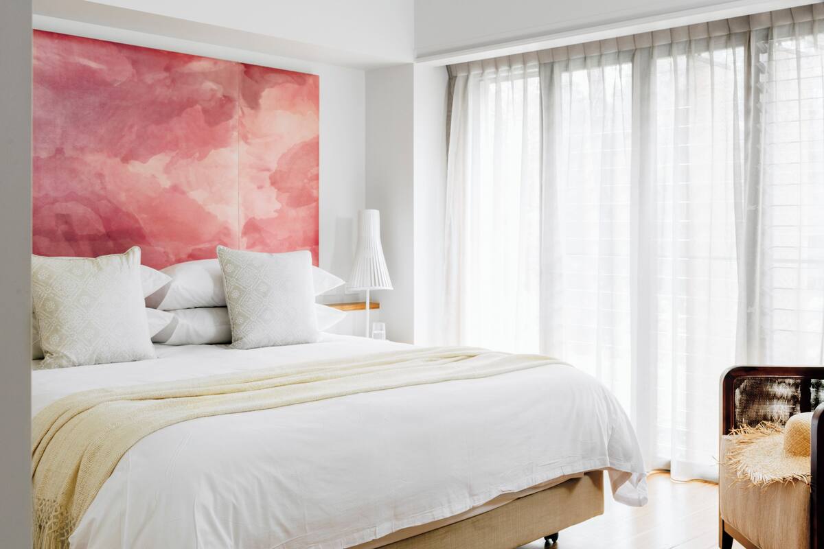 A modern bedroom with a large bed, a wall art piece, a bedside lamp, and a window with sheer curtains.