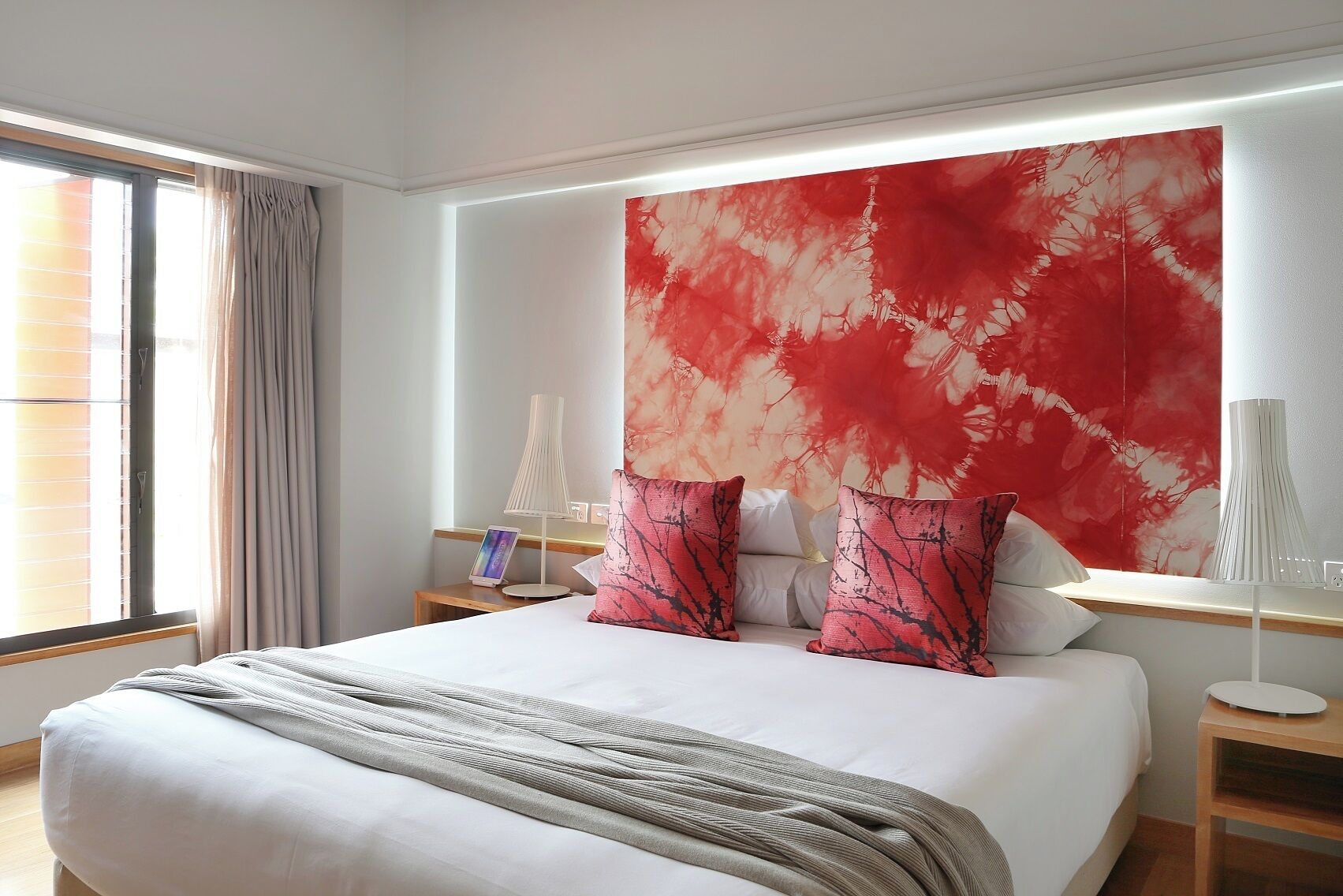 A modern hotel room with a large bed, a bedside table, a nightstand, a lamp, and a large abstract painting on the wall.