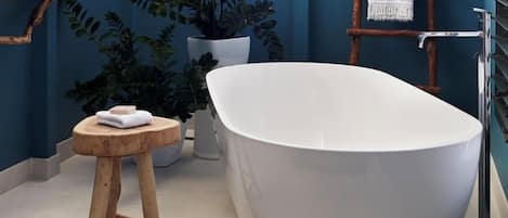 A modern bathroom with a freestanding bathtub, a wooden stool, a potted plant, and a towel rack.