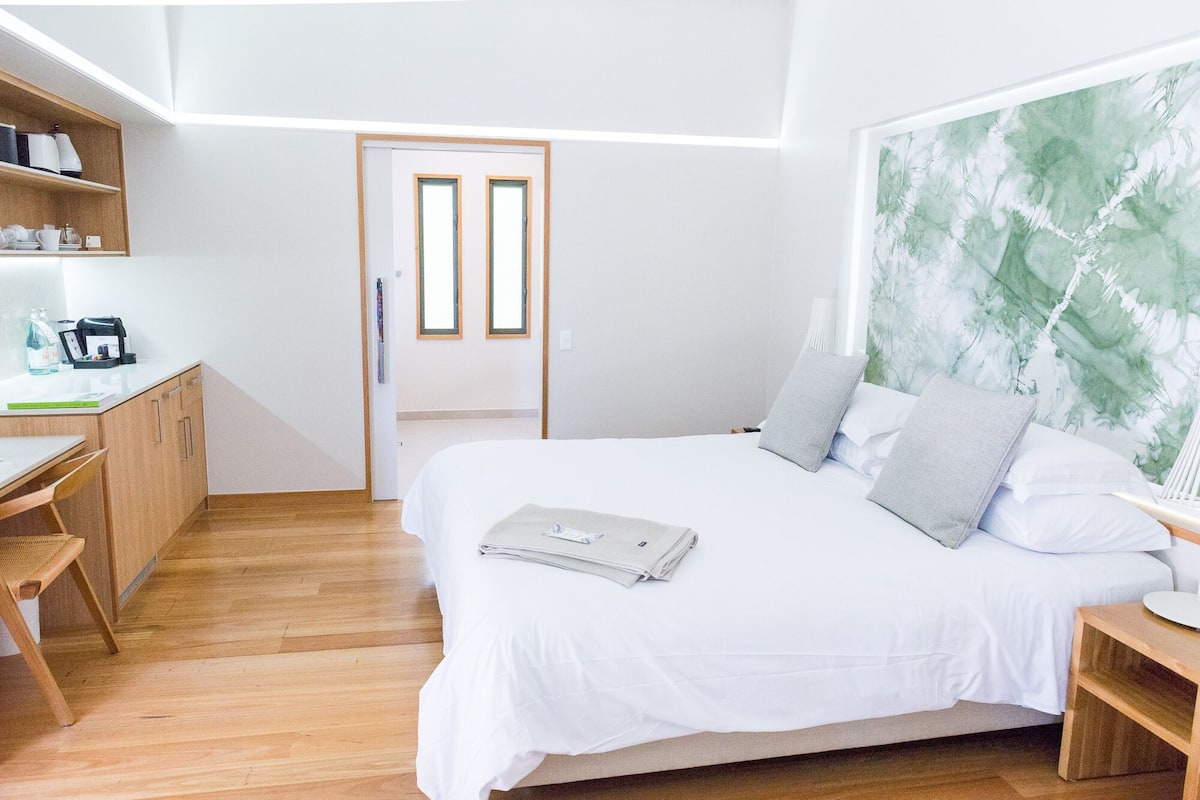 A modern bedroom with a wooden floor, a bed with white bedding, a desk with a coffee machine, and a large abstract painting on the wall.