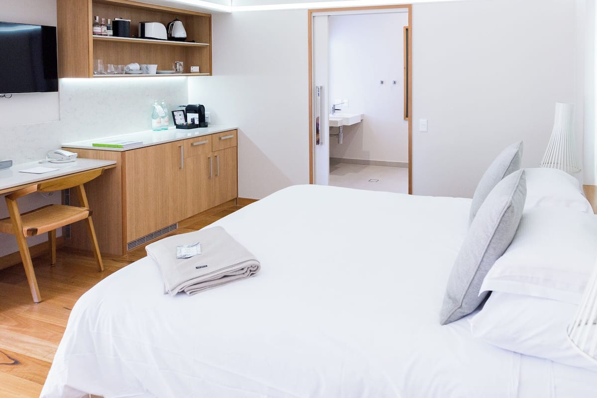 A hotel room with a bed, a desk with a chair, a television, a shelf with kitchen items, and a bathroom visible through an open door.