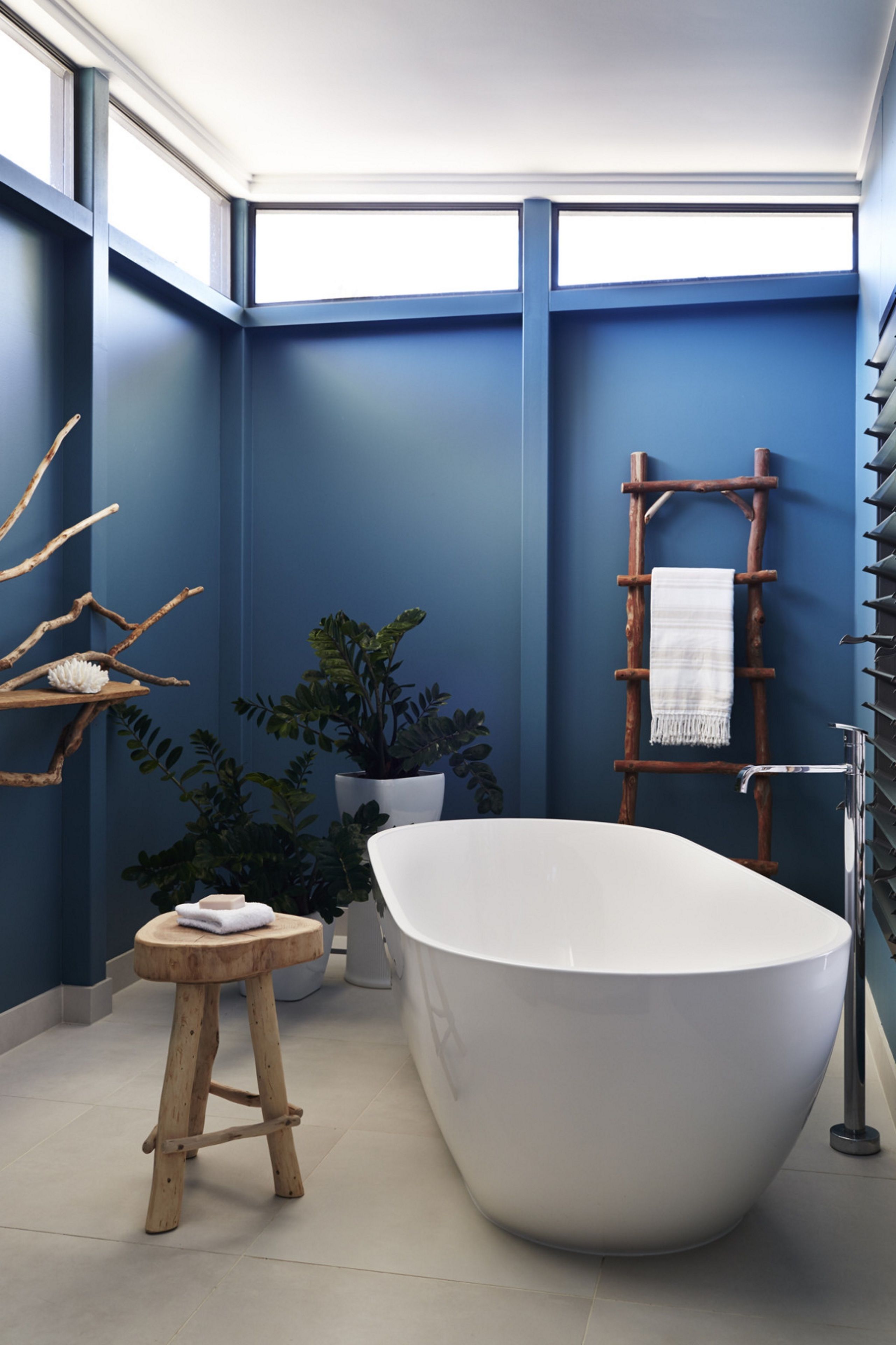 A modern bathroom with a freestanding bathtub, wooden stool, potted plants, and a towel rack.
