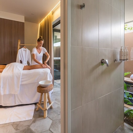 A spa room with a massage table, a wooden stool, a sink, and a small garden area visible through the glass wall.