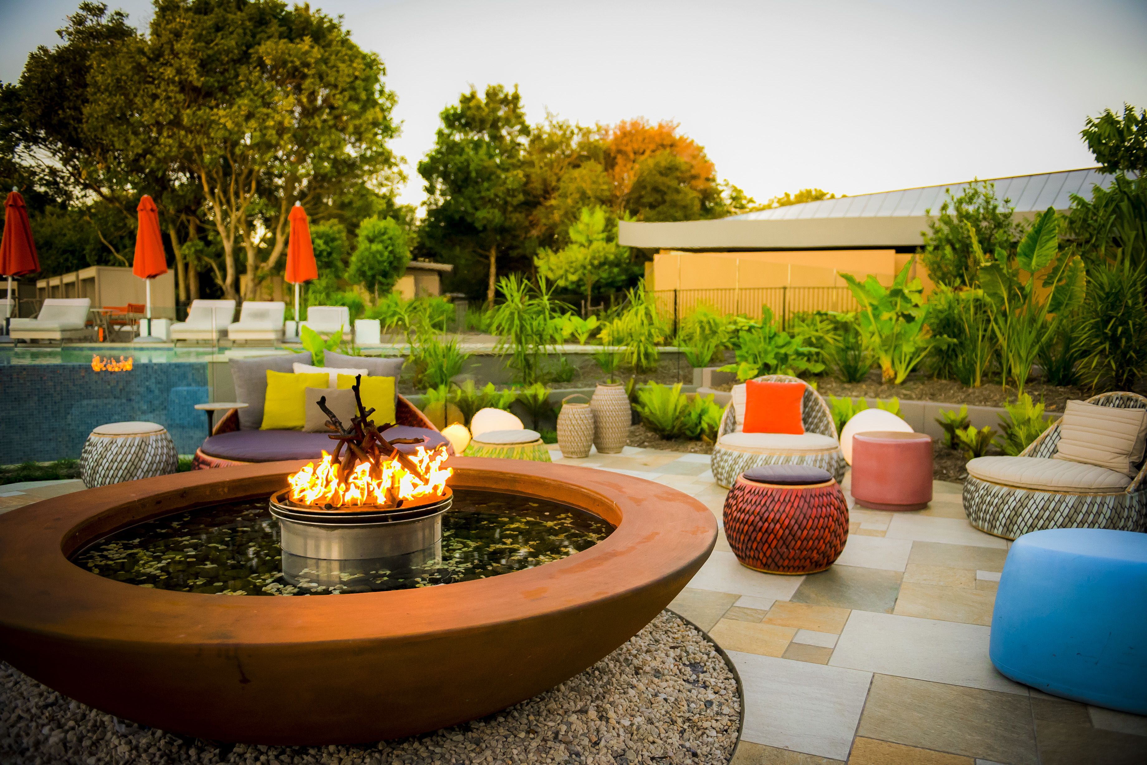 A fire pit surrounded by outdoor seating, a pool, and lush greenery.
