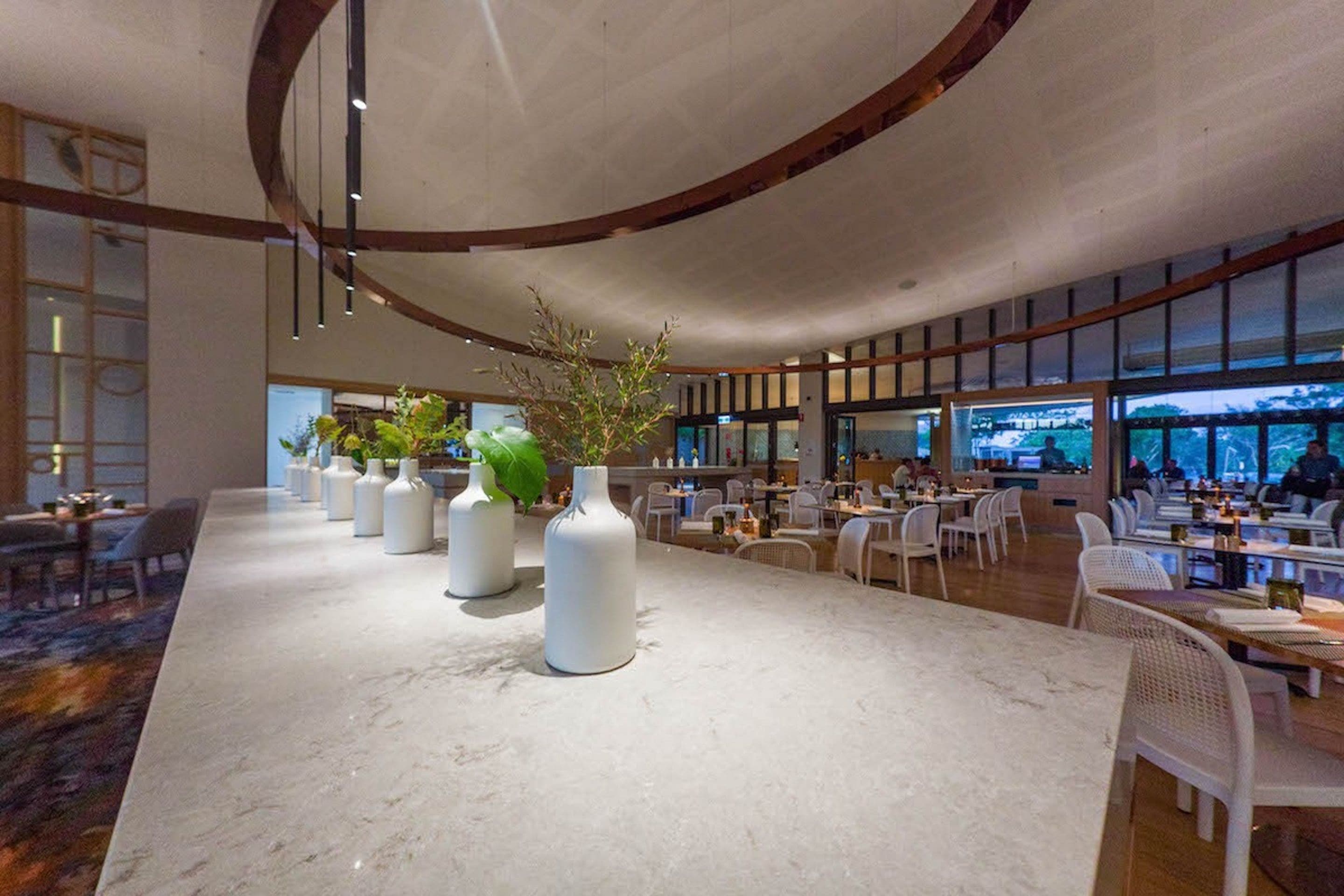 A spacious indoor dining area with a long marble counter, white vases with plants, and a modern ceiling design.