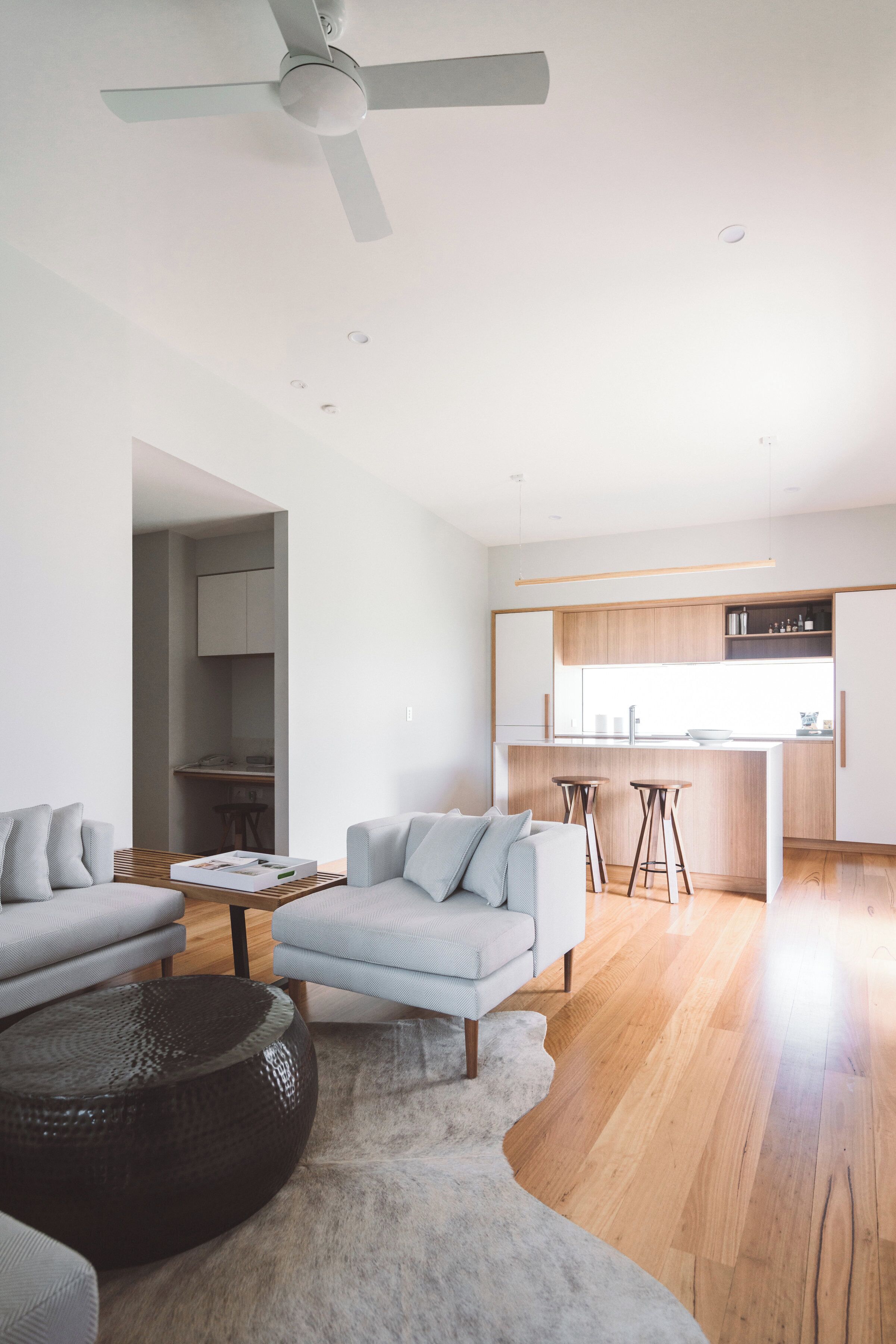 A modern living room with a sofa, coffee table, and a rug. There is a kitchen area with wooden cabinets and a bar stool.