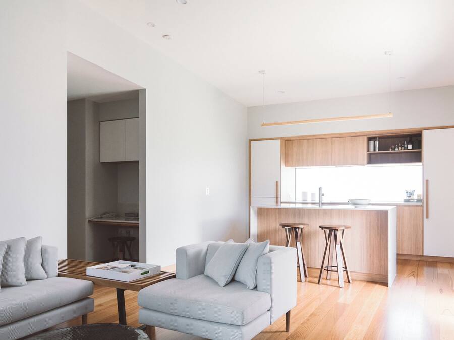 A modern living room with a sofa, coffee table, and a rug. There is a kitchen area with wooden cabinets and a bar stool.