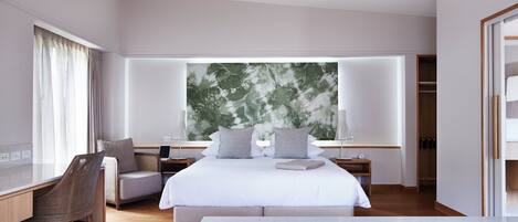 A modern hotel room with a large bed, a desk, a chair, and a view of greenery through the window.