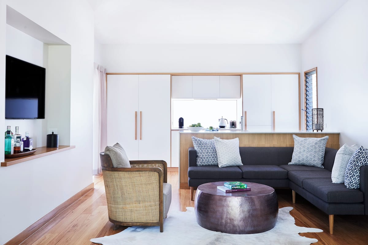 A modern living room with a sofa, a coffee table, and a wicker chair. A kitchen is visible in the background with a window above the sink.