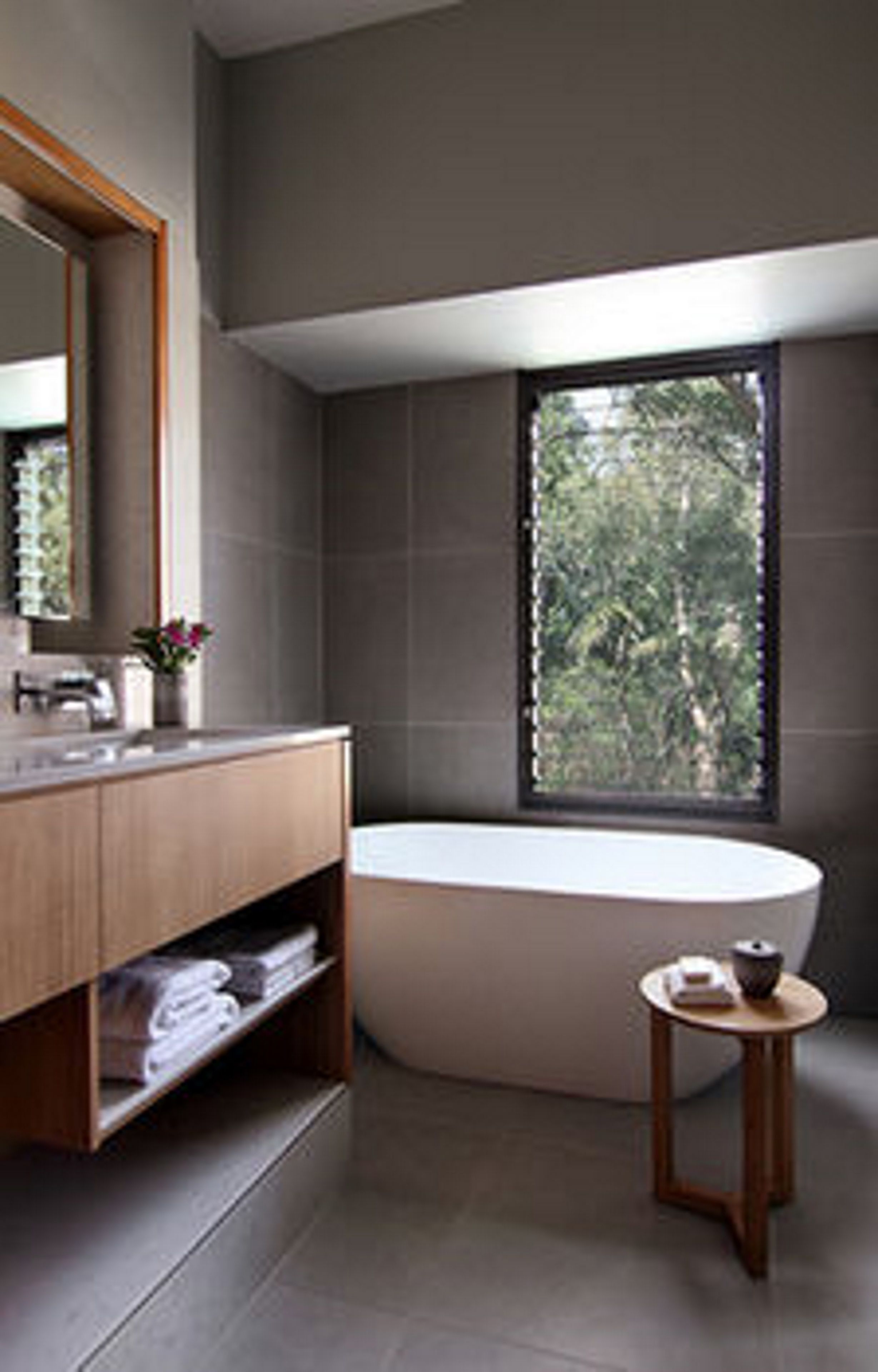 A modern bathroom with a large bathtub, a wooden vanity, a small round table, and a window with a view of greenery.