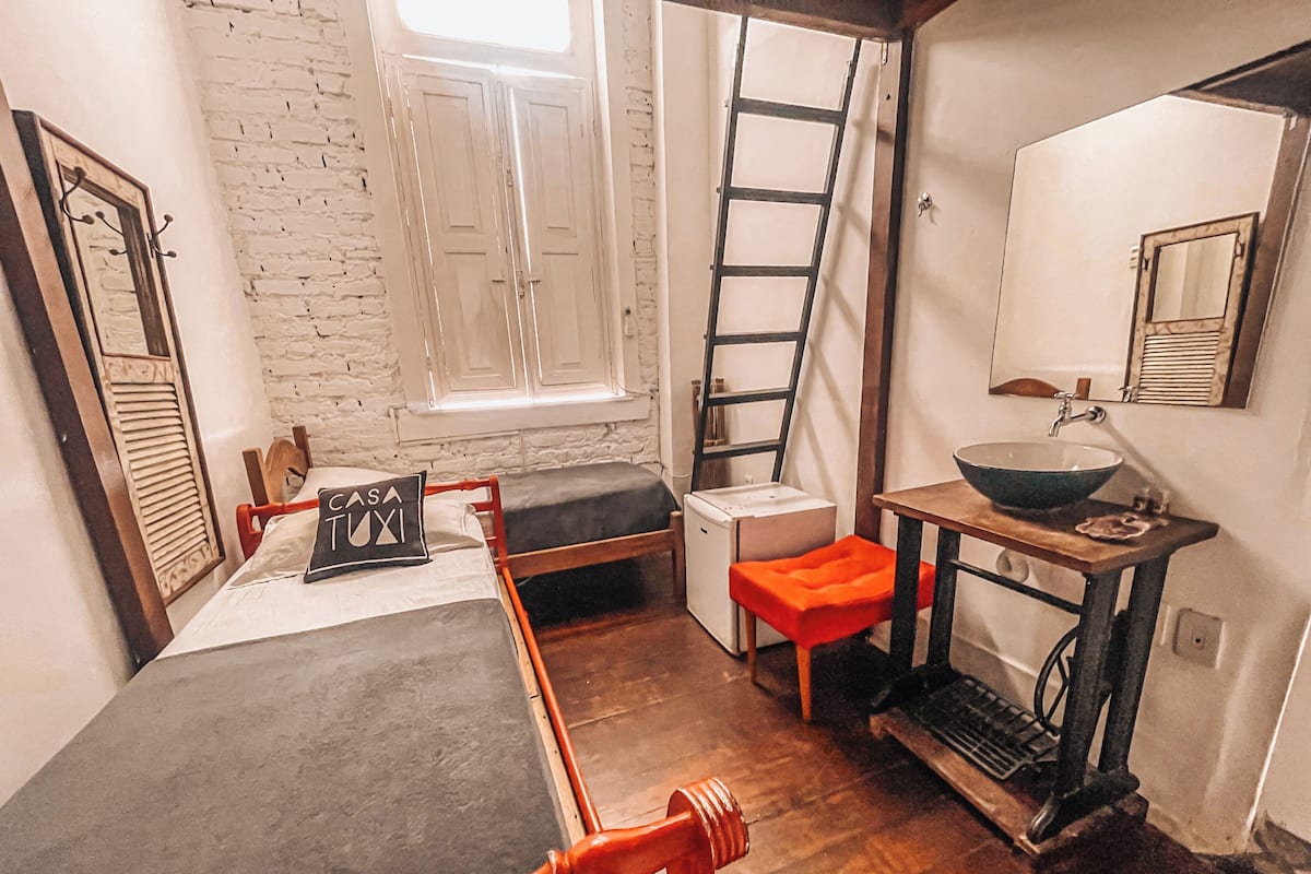 A compact room with a bed, a small table with a sink, a ladder, and a window with shutters.