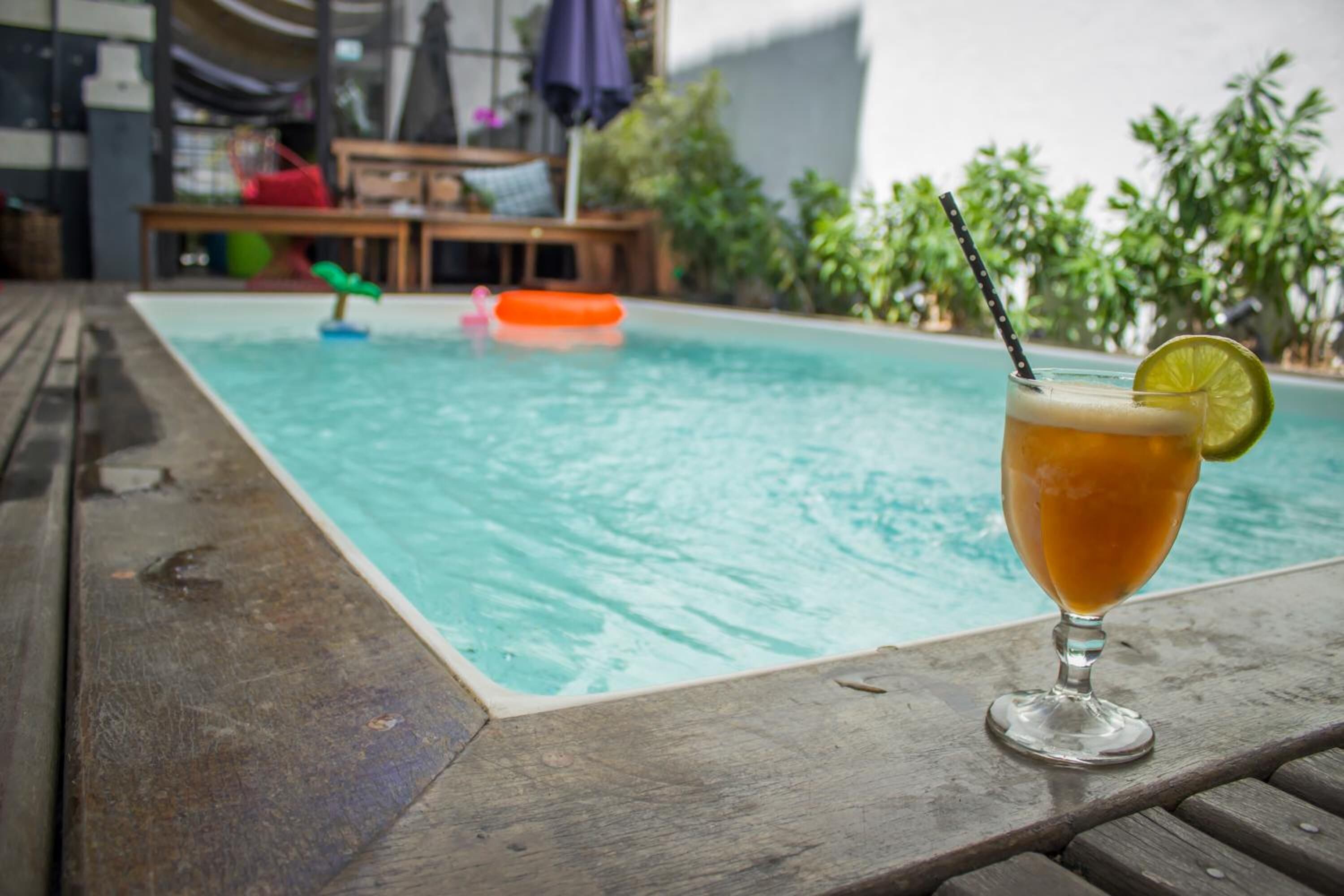 Una cerveza con una rodaja de lima sobre una mesa de madera junto a una piscina con juguetes flotantes.