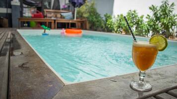 Una cerveza con una rodaja de lima sobre una mesa de madera junto a una piscina con juguetes flotantes.