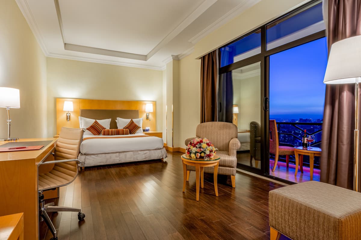 A hotel room with a bed, desk, and chair, a small table with a flower arrangement, and a balcony with a view.