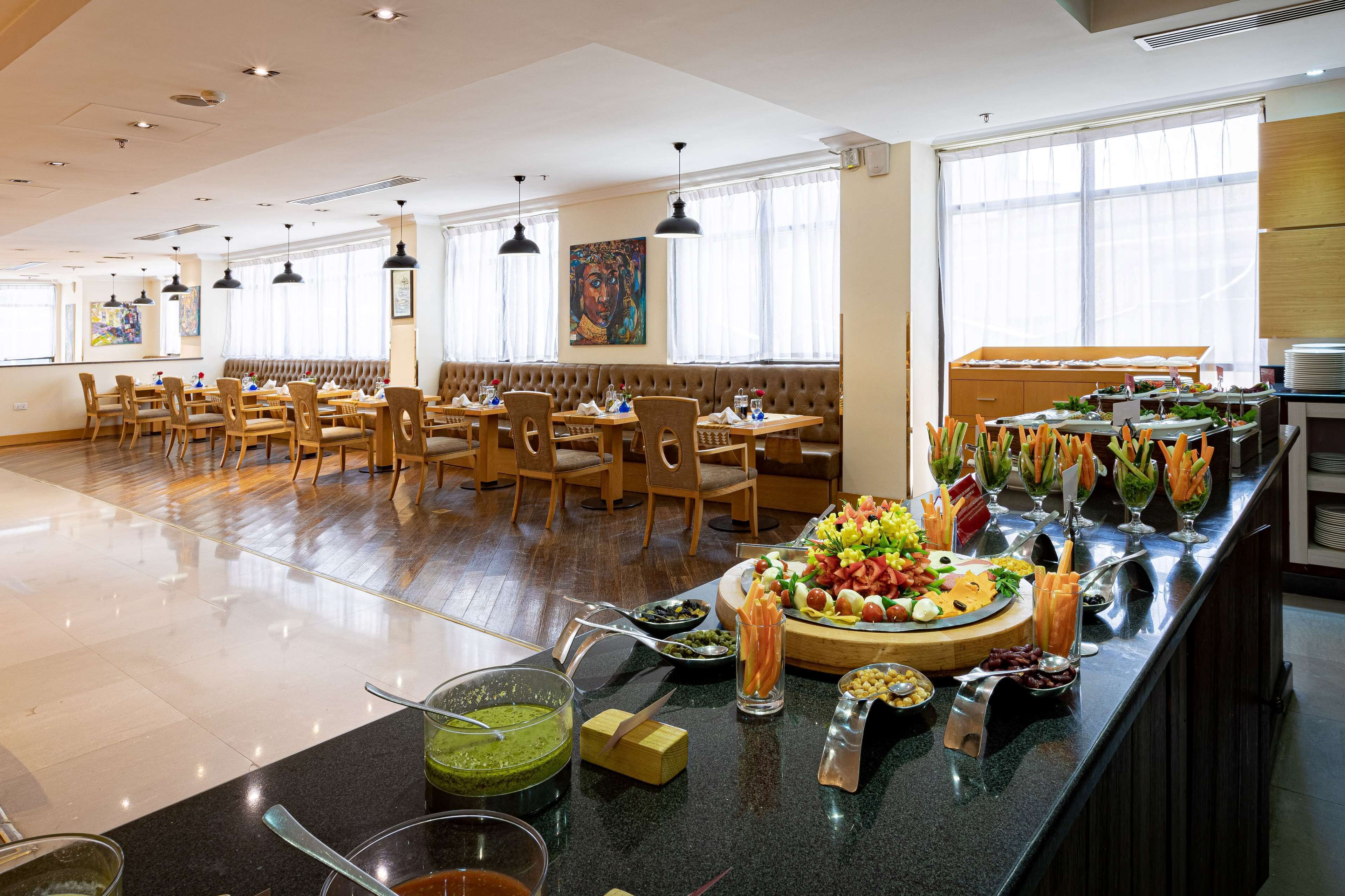 A spacious dining area with wooden tables and chairs, a buffet with various food items, and large windows providing natural light.