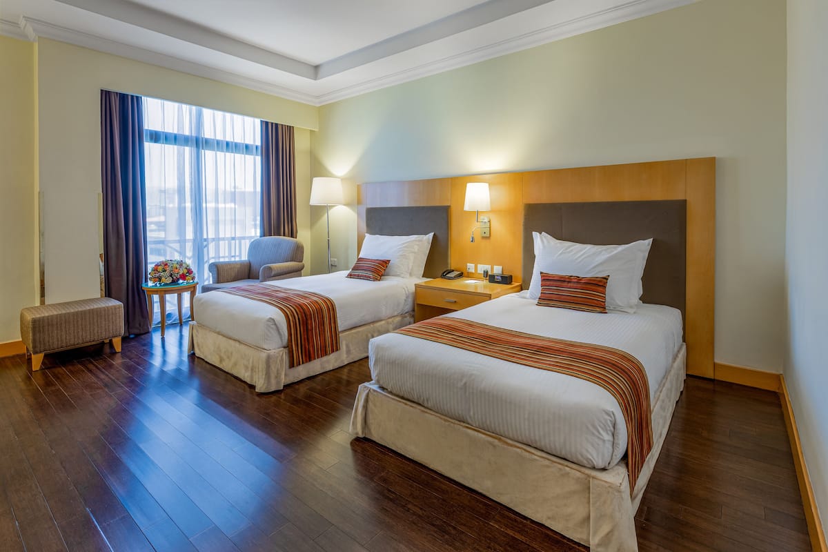 A hotel room with two beds, a bench, a chair, a table with a lamp, and a window with curtains.