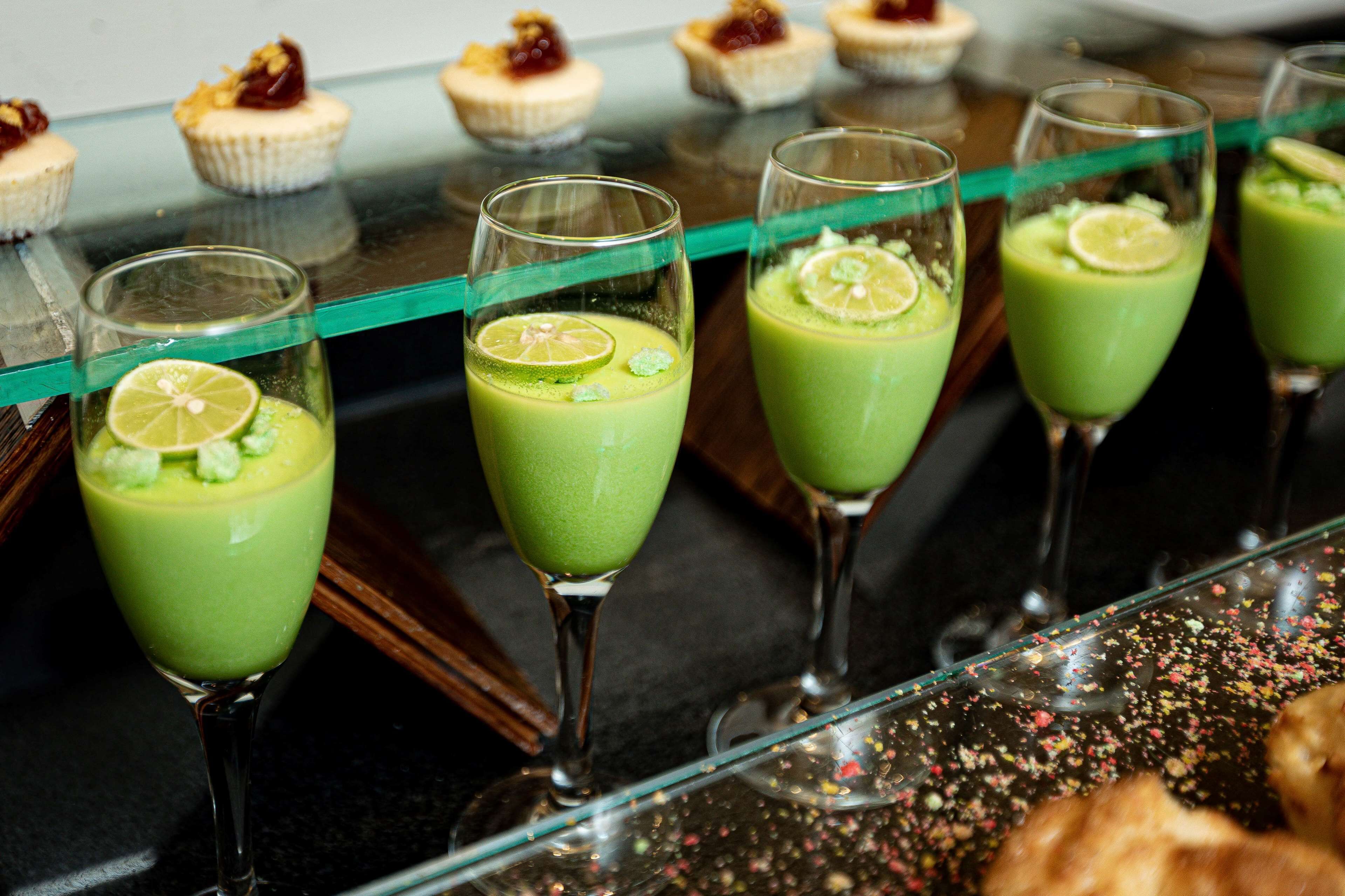 A display of green smoothies in glasses, with a glass shelf above containing small desserts.
