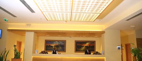 Un mostrador de recepción con dos recepcionistas, una lámpara de techo grande y dos cuadros en la pared.