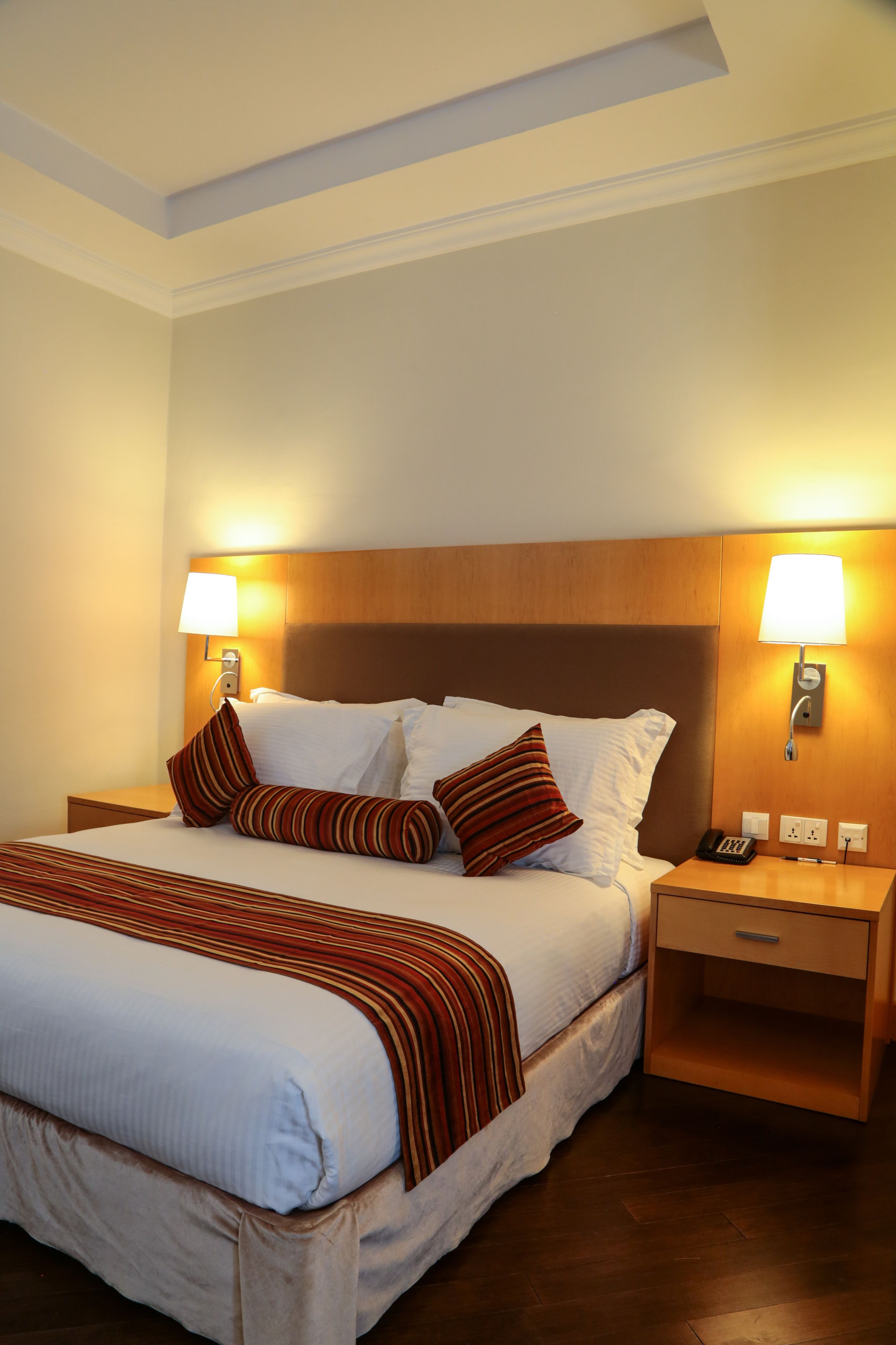 A hotel room with a large bed, two bedside tables, a wooden headboard, and two wall-mounted lamps.