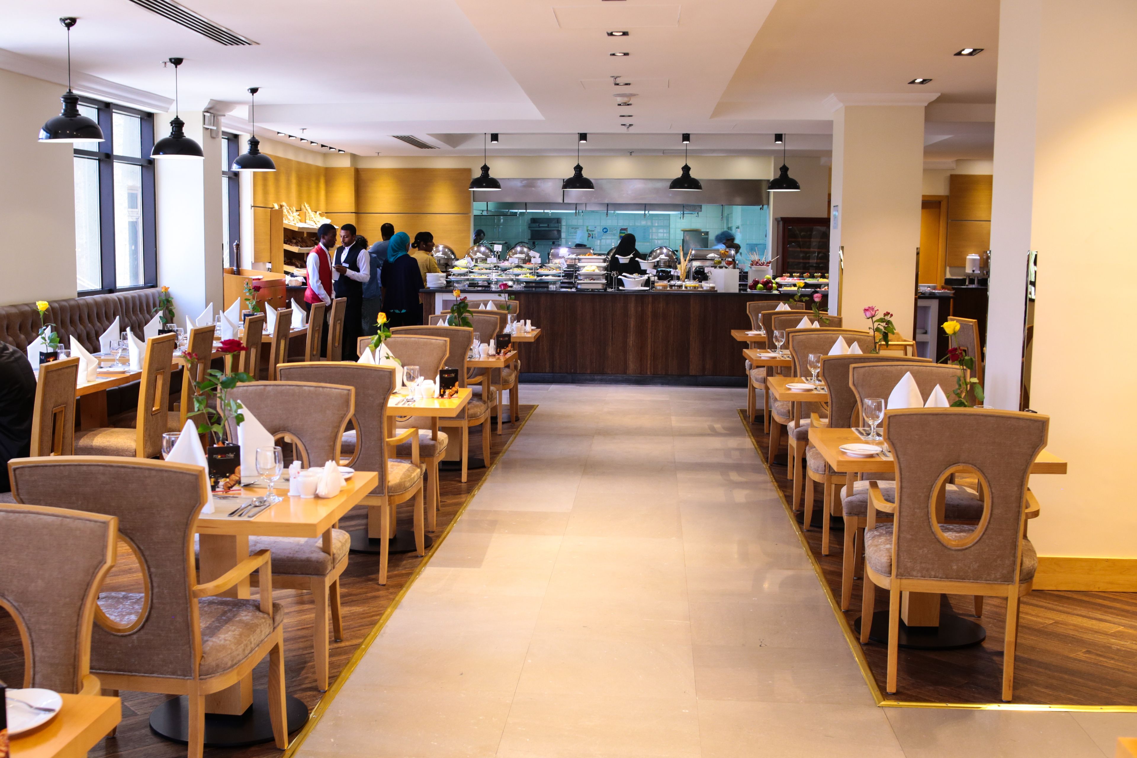 Un restaurant amb diverses taules i caires per dinar, una barra amb personal i una cuina al fons.